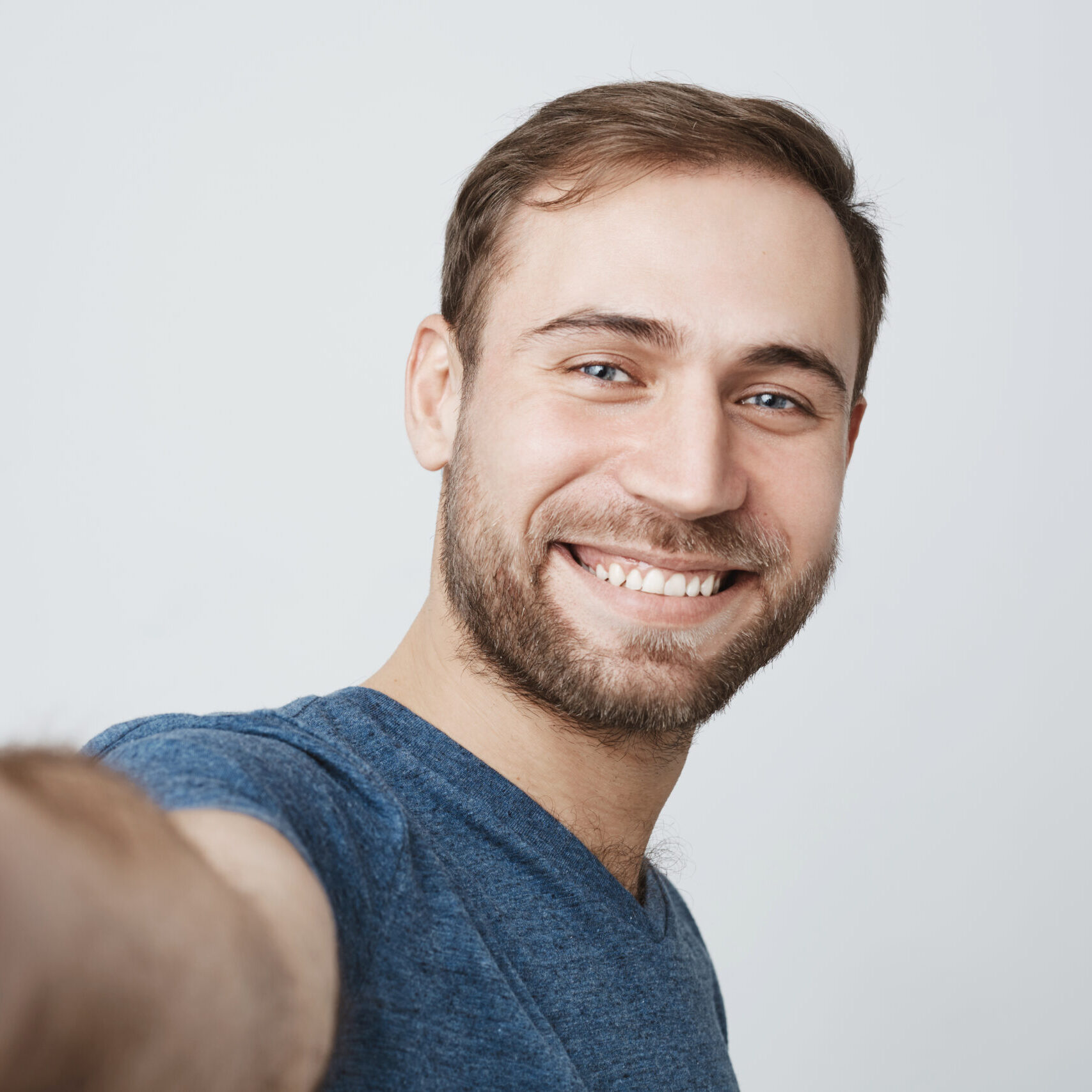 Close-up portrait of smiling bearded face of good-looking guy in blue shirt, looking at the camera, relaxing indoors, posing against gray background. Positive emotions and feelings.